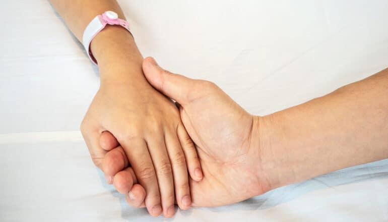 A parent holds their sick child's hand while they lay in a hospital bed.