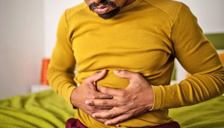 A young man clutches his stomach in pain while sitting on a bed.