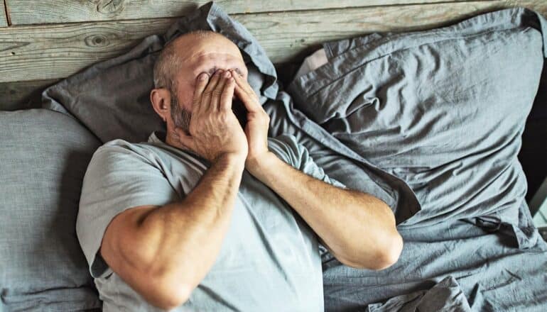 A man covers his face with both hands as he wakes up in bed.