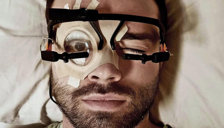 A man sleeps with one eye closed and the other held open by a device strapped to his face.