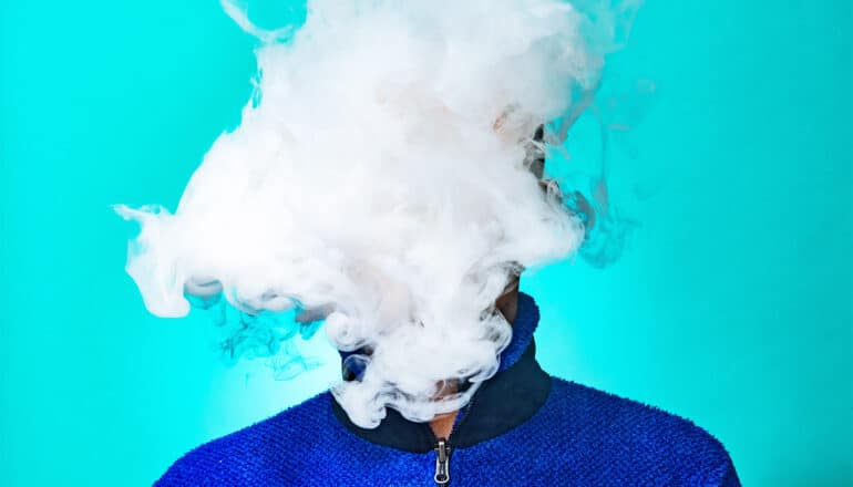 A cloud of vapor covers a person's face as they stand in front of a bright blue background.